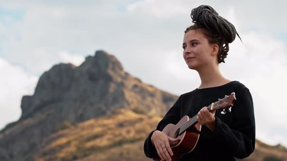 Young Woman with Dreadlocks Playing Ukulele in Mountains and Looking in the Camera alt