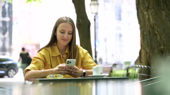 Portrait of Young Woman Resting in Cafe Outdoors and Using Smartphone for Texting alt