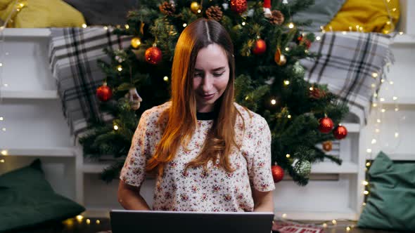 Cute girl with laptop working by the christmas tree in the evening