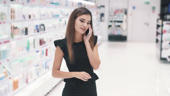 Front View of Young Caucasian Woman Talks By Smartphone in Cosmetics Store alt