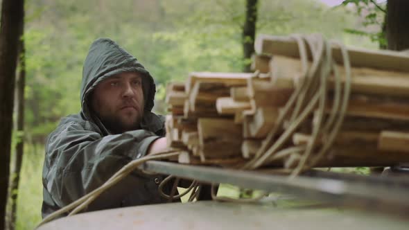 Slow Motion Of Casual Strong Man Works with Firewood and Rope Near Car in Wild Deciduous Forest. alt