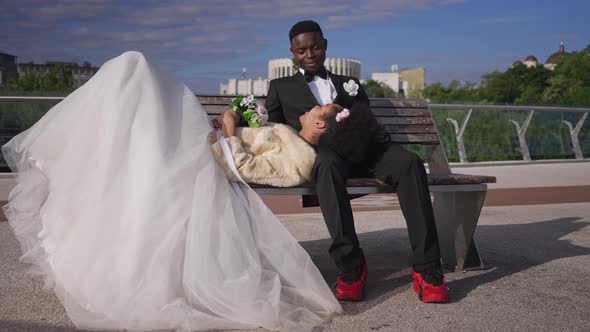 Wide Shot of Smiling Happy Groom Sitting on Bench with Bride Lying on Knees alt