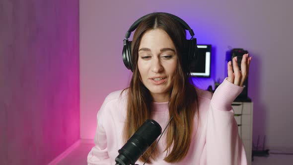 Happy Young Woman of Headphones Looking at Camera and Talking Smiling While Sitting at Desk in Home alt