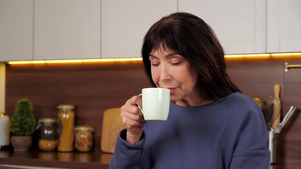 Aged Woman Drinking Delicious Warm Tea Coffee From Cup in Home Kitchen alt