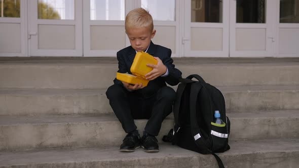 A Schoolboy Opens a Lunch Box with Food Sitting on the Steps of the School alt