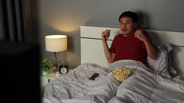 shocked young man is watching horror movie TV on a bed at night