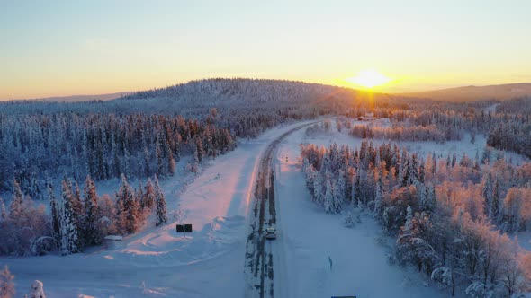 Aerial view above vehicle joining snowy remote road heading towards woodland mountain sunrise alt