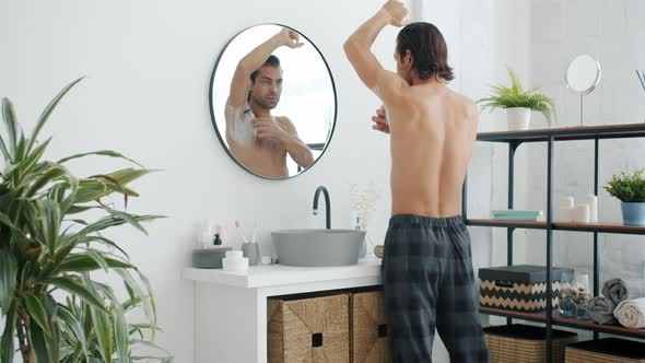 Back View of Young Man in Pajamas Pants Shaving Armpits with Razor in Bathroom alt