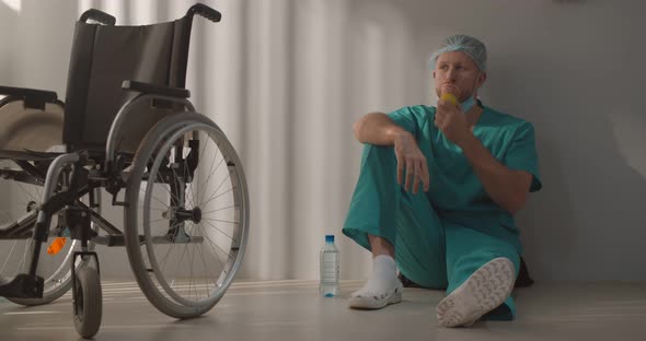 Young Male Nurse or Surgeon in Scrubs Sitting on Hospital Floor and Eating Apple alt