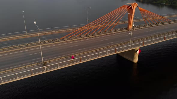 Aerial Backside View: Two People Ride Bicycles on Bridge Across River During Sunset alt