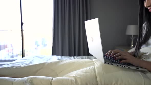 A young woman sitting on a bed in room and works on their laptop