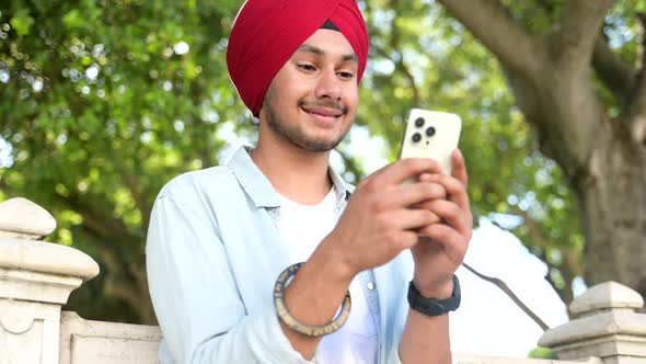 Carefree Young Indian Guy in Casual Shirt and Traditional Headdress Using Smartphone Outdoors