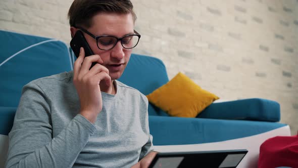 Man Talking on Phone While Is Working on Laptop Sitting on Bean Bag at Home alt
