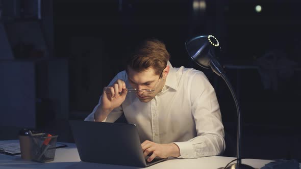 A Tired Man is Looking at a Financial Report on a Computer in the Office at Night