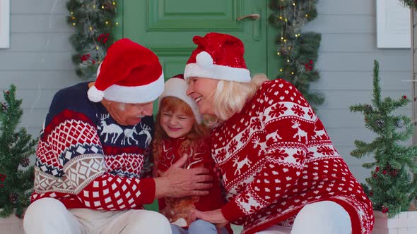 Senior Grandparents with Granddaughter in Santa Claus Hat Celebrating Christmas Near Decorated House alt