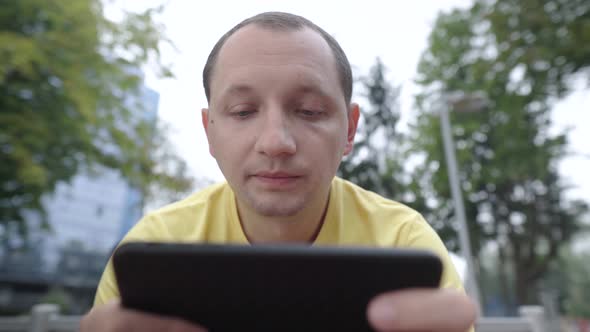 Portrait of Young Man in a Yellow Tshirt Chews Gum and Looks Intently at His Smartphone