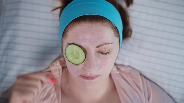 Brunette Woman Lies with a Cosmetic Mask and Eats a Cucumber