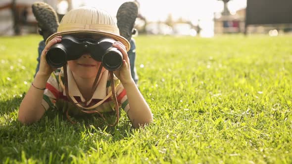Child Wearing Pith Helmet is Playing in Summer Day with Binoculars Lying Down on the Grass at Town
