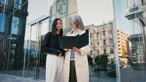 Asian Mature Woman Holding Folder and Explaining Details of New Project to Young Colleague Pointing alt