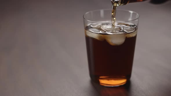 Slow Motion Man Pour Cola Over Ice Cubes Into Tumbler Glass on Walnut Table with Copy Space alt
