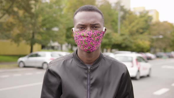 A Young Black Man in a Face Mask Points at the Camera and Nods in a Street in an Urban Area alt