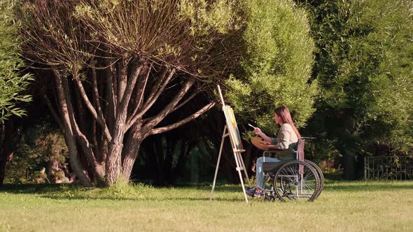 A Disabled Woman in a Wheelchair Paints a Picture in the Park Near a Tree alt