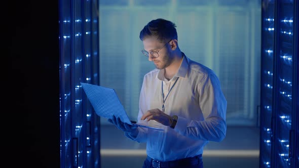 Male Network Engineer Doing a System Check Standing in the Server Room with His Laptop alt