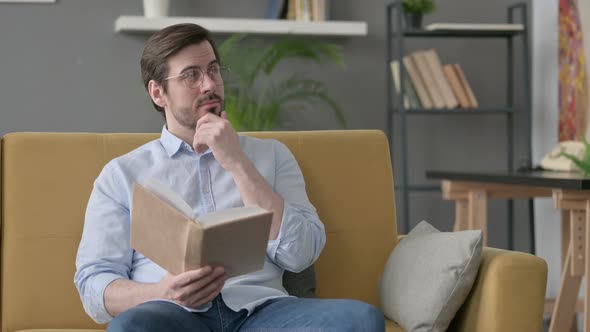 Young Man Thinking While Reading Book on Sofa alt