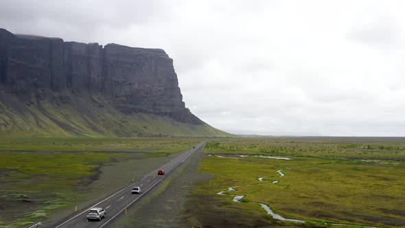 Three cars driving on Ring Road in Iceland by a mountain with drone video following behind. alt