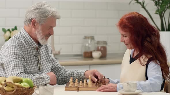 Cheerful Senior Couple 6070 Years Old Play Chess at Home Sitting at the Table in the Kitchen alt