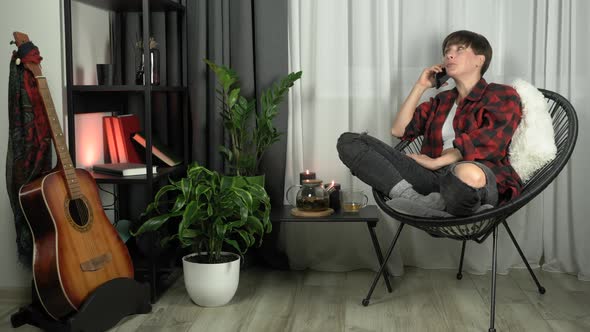 Woman is talking by mobile phone sitting in cozy chair in living room. alt