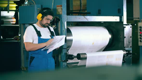 Male Worker Is Taking Notes Next To a Working Printing Press alt