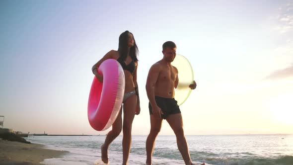 Happy Young Couple Walking on the Beach with Rubber Rings During Sunset Slow Motion alt