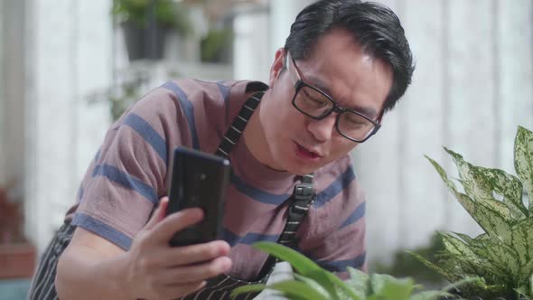 Asian Man Waving Hands And Talking While Live Stream Selling Plants By Smartphone At Home alt