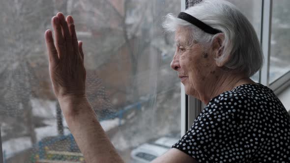 Old Elderly Grandmother Woman Looks Calm Out Of Window With Hand On The Glass alt