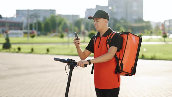 Asian Courier Food Delivery With Red Thermal Backpack With Electric Scooter Uses Smartphone Navigate