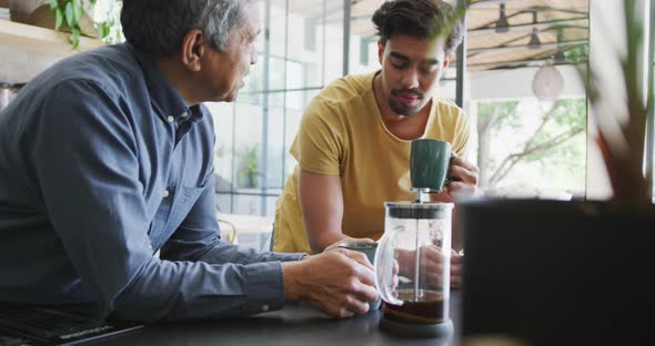 Man talking to young son drinking coffee from mug while leaning on kitchen counter at home alt
