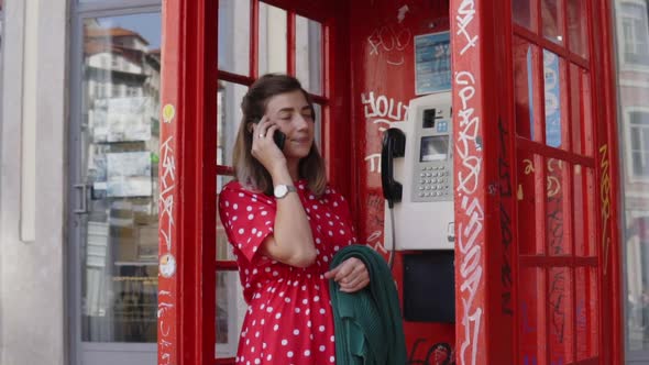 Young Woman, Student Is Talking on Mobile Phone in Telephone Box alt