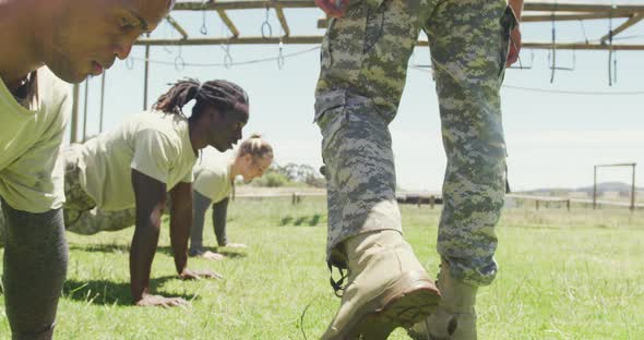 Legs of instructor walking past diverse soldiers doing press ups at army obstacle course in sun alt
