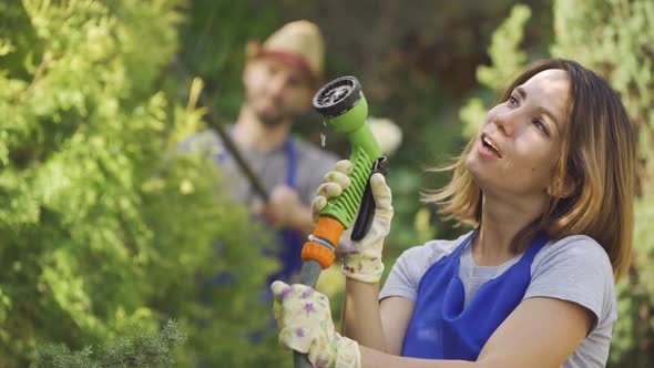 Portrait of Attractive Caucasian Woman Playing with Water Hose in the Foreground While Man Cutting alt