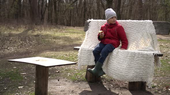 a Little Boy in a Red Jacket in the Park Sits on a Bench and Eats an Apple