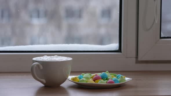 A Large Mug of Coffee with Milk or Mochacino and Colorful Sweets in a Plate on the Windowsill in