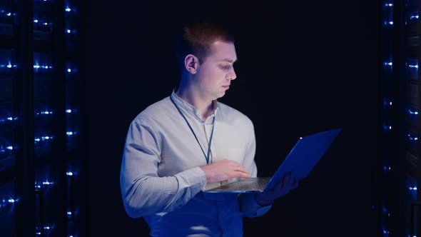 Young Man Holds Device in Hand and Looks at Screen Inspects Equipment or Hardware Rack alt