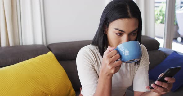 Gender fluid male drinking coffee and using smartphone while sitting on the couch at home alt