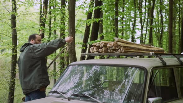 Slow Motion Of Casual Strong Man Works with Firewood and Rope Near Car in Wild Deciduous Forest. alt