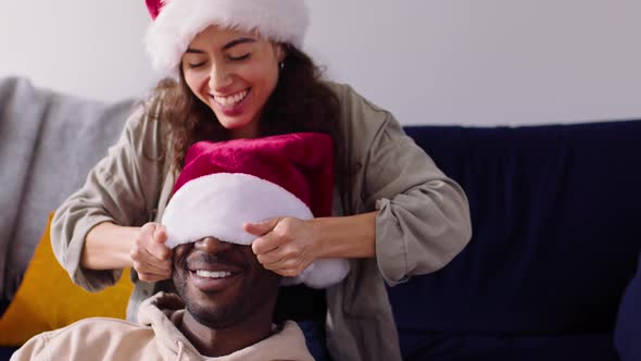 Woman Pulling Santa Hat Over Mans Face As Couple Prepare For Christmas At Home