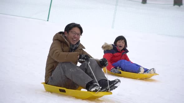 Asian Father And His Son Sliding On Sled Under Snow Fall alt