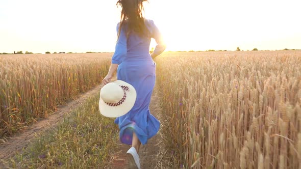 Beautiful Woman in a Blue Dress and Hat Runs Through a Wheat Field at Sunset alt