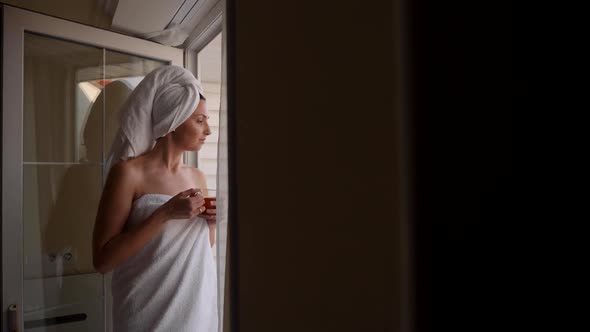 Woman in a Towel After a Bath is Standing Next to an Open Door on the Balcony alt