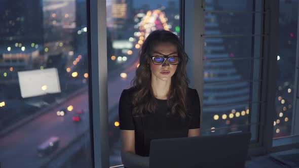 Work Late at Night in the Office. Young Woman in Business Suit and Glasses Works on a Laptop on the alt